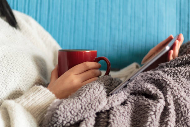 Woman lying on sofa with blanket watching a movie on tablet and holding a mug